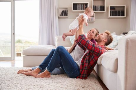 couple sitting on floor of living room with husband holding baby up in air with two arms and wife smiling as she watches 