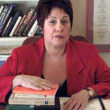 Dr. Phyllis chesler wearing red jacket sitting at desk with right hand on a pile of books and library shelf behind her
