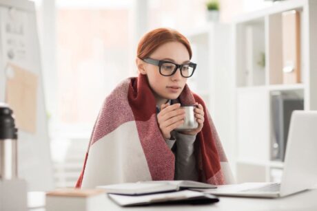 young woman wearing glasses holding cup of tea wrapped in plaid blanket reading about health issues