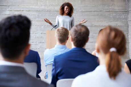 woman standing a podium speaking a training program to men and women in business